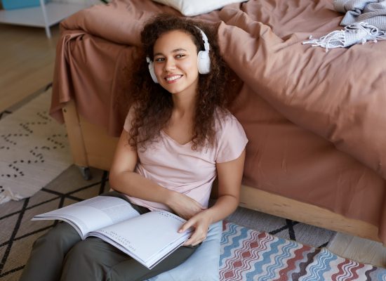 portrait-positive-young-curly-mulatto-lady-room-dressed-pajamas-enjoying-his-favorite-music-headphones-reading-new-magazine-about-art-smiling-looks-away